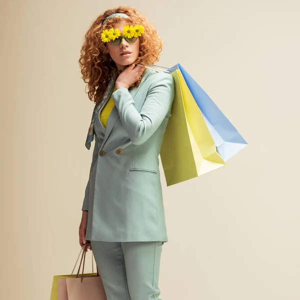 Приваблива руда дівчина в костюмі і сонцезахисних окулярах з квітами тримає сумки для покупок ізольовані на бежевому — стокове фото