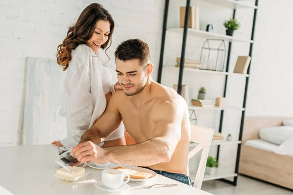 Соблазнительная девушка в белой рубашке сидит на кухонном столе, пока сексуальный парень завтракает — стоковое фото