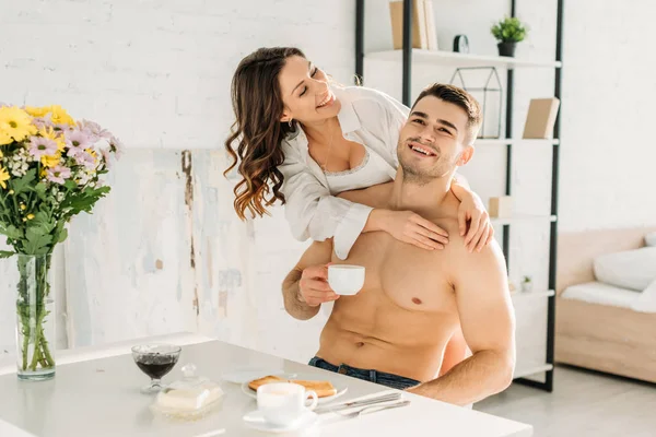 Alegre sexy chica abrazando feliz sin camisa novio beber café mientras desayunando - foto de stock