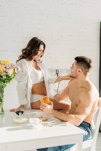 Menina sedutora em camisa branca e lingerie sentado na mesa da cozinha e tocando namorado sexy tomando café da manhã — Fotografia de Stock