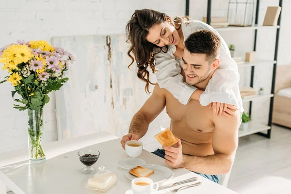 Sexy girl in white shirt hugging happy shirtless boyfriend having breakfast in kitchen — Stock Photo
