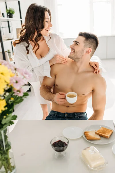 Feliz pareja mirándose mientras el hombre toma café en la mesa de la cocina con el desayuno - foto de stock