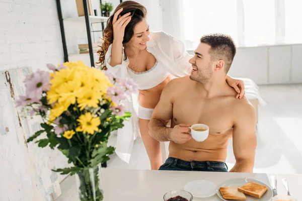 Sexy ragazza in camicia bianca e lingerie toccare felice uomo bere caffè vicino vaso con fiori — Foto stock