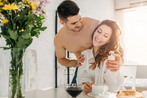 Сексуальный мужчина без рубашки обнимает счастливую девушку сидя за кухонным столом рядом с вазой с цветами — стоковое фото