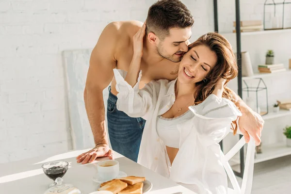 Красивий без сорочки чоловік цілує усміхнену дівчину, торкаючись його обличчя, сидячи біля сніданку — стокове фото