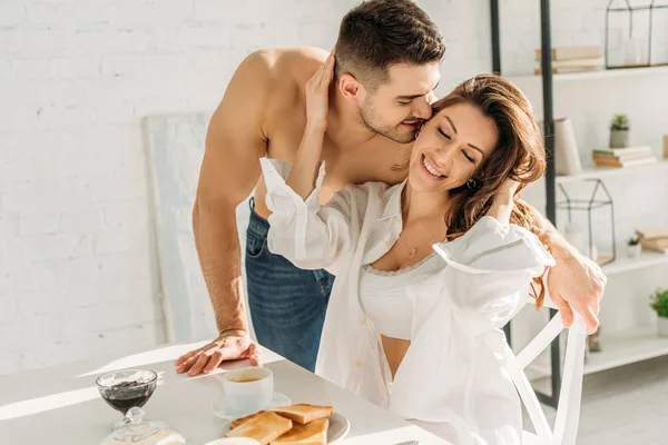 Guapo sin camisa hombre besar sonriente chica tocando su cara mientras está sentado cerca del desayuno - foto de stock