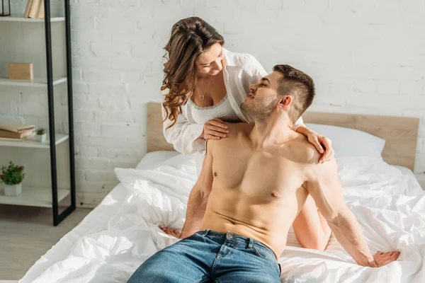 Sensual menina em branco camisa e bralete olhando sem camisa, sexy homem sentado na cama em jeans — Fotografia de Stock