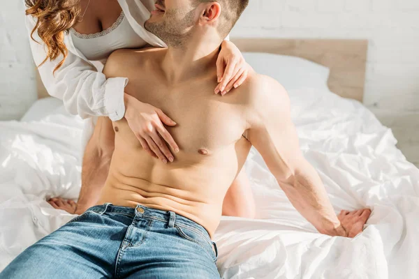 Cropped view of sexy girl in white shirt and bralette hugging shirtless man sitting on bed in jeans — Stock Photo