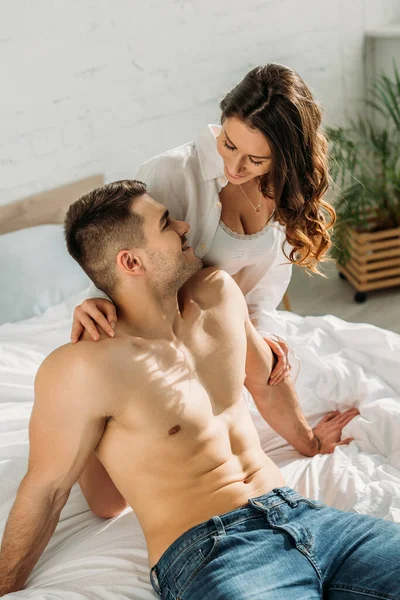 Чуттєва дівчина і сексуальний без сорочки хлопець посміхається один одному, сидячи на ліжку — Stock Photo