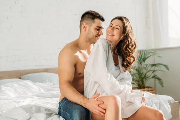 Улыбающийся мужчина без рубашки прикасается к ноге сексуальной, веселой девушки, сидя на кровати — стоковое фото