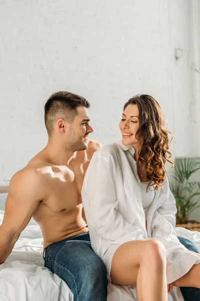 Hombre sin camisa en jeans y chica sexy en camisa blanca sonriendo el uno al otro mientras se sienta en la cama - foto de stock