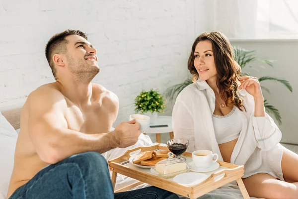 Homme torse nu souriant tenant tasse de café et levant les yeux tout en étant assis près de petite amie sexy et plateau de lit avec délicieux petit déjeuner — Photo de stock