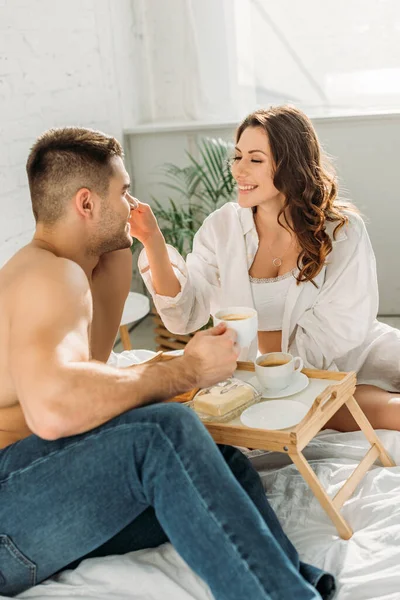 Happy, sexy girl touching face of shirtless boyfriend near bed tray with delicious breakfast — Stock Photo