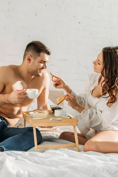 Attractive girl giving toast to sexy boyfriend holding cup of coffee near bed tray with tasty breakfast — Stock Photo