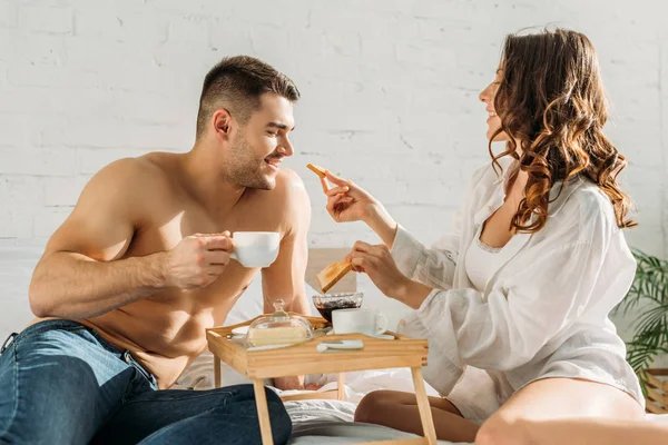Smiling girl giving toast to shirtless boyfriend holding cup of coffee near bed tray with delicious breakfast — Stock Photo