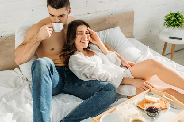 Bell'uomo senza camicia che beve caffè vicino alla fidanzata allegra e al vassoio del letto con una gustosa colazione — Foto stock