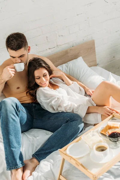 Bel homme torse nu buvant du café et femme sexy souriant près du plateau de lit avec petit déjeuner savoureux — Photo de stock