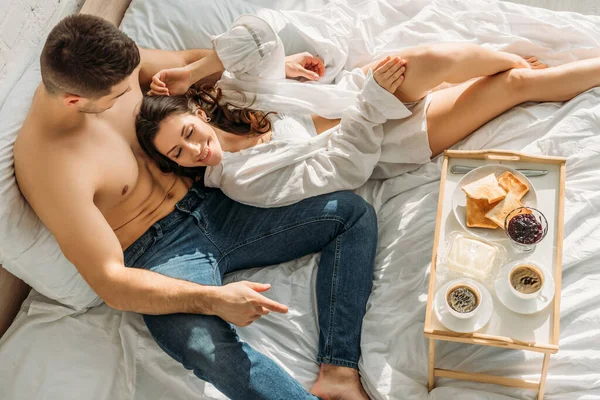 Draufsicht auf einen hemdlosen Mann in Jeans und ein sexy Mädchen im weißen Hemd, das neben dem Bettlaken mit leckerem Frühstück liegt — Stockfoto