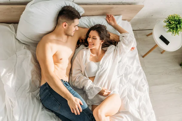 Вид зверху усміхненого безсорочного чоловіка в джинсах і сексуальна, весела дівчина в білій сорочці, що лежить у ліжку разом — стокове фото