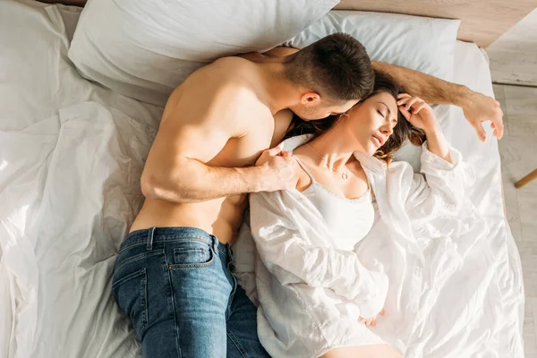 Vista superior do homem sem camisa em jeans abraçando namorada deitada na cama com os olhos fechados — Fotografia de Stock