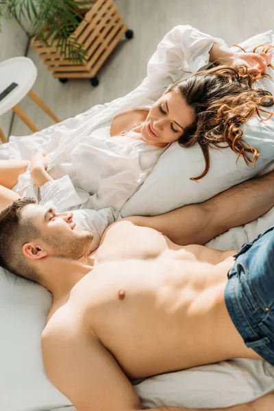 Vista aérea de sexy pareja joven mirándose el uno al otro y sonriendo mientras está acostado en la cama - foto de stock