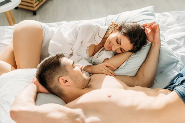 Sexy sin camisa hombre tocando la cabeza de feliz novia mientras está acostado en la cama juntos - foto de stock
