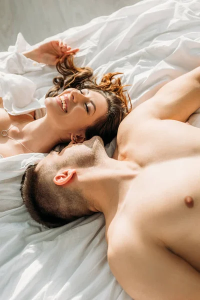 Vista aérea de hombre sin camisa sexy acostado en la cama cerca de sensual, novia feliz - foto de stock