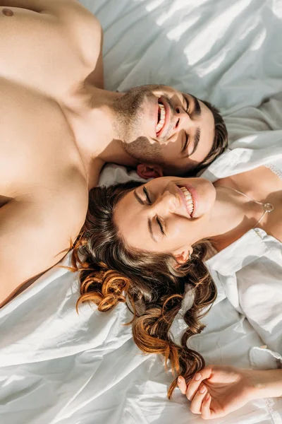 Top view of cheerful, sexy couple smiling with closed eyes while lying in bed together — Stock Photo