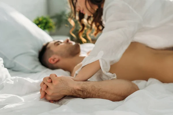 Вибірковий фокус спокусливої дівчини тримає руки з сексуальним чоловіком, лежачи на ньому в ліжку — стокове фото