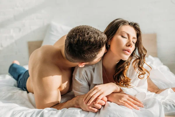 Young shirtless man kissing passionate girlfriend lying in bed with closed eyes — Stock Photo