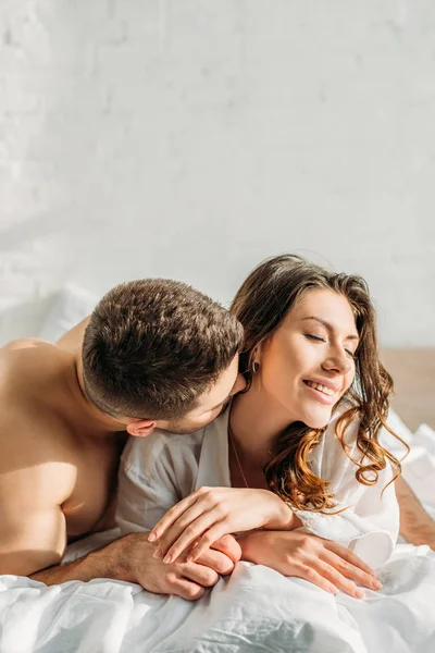 Joven sin camisa hombre besar seductora novia sonriendo en la cama con los ojos cerrados - foto de stock