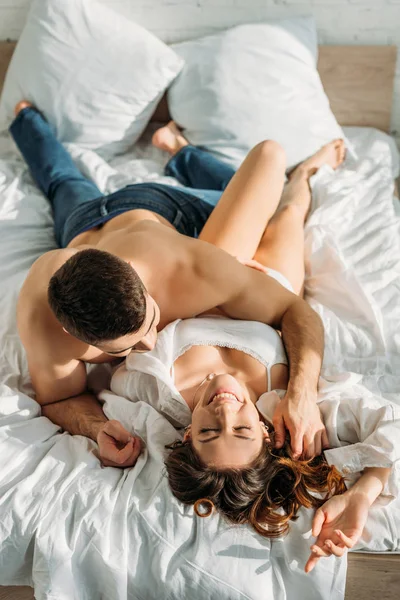 Над головой вид сексуального мужчины без рубашки обнимающего улыбающуюся девушку, лежащего в постели вместе — стоковое фото
