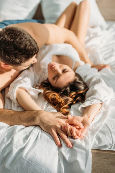 Selective focus of young man holding hand of sexy girl while lying in bed together, overhead view — Stock Photo