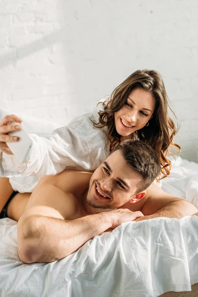 Молодая веселая пара делает селфи на смартфоне, лежа в постели вместе — стоковое фото