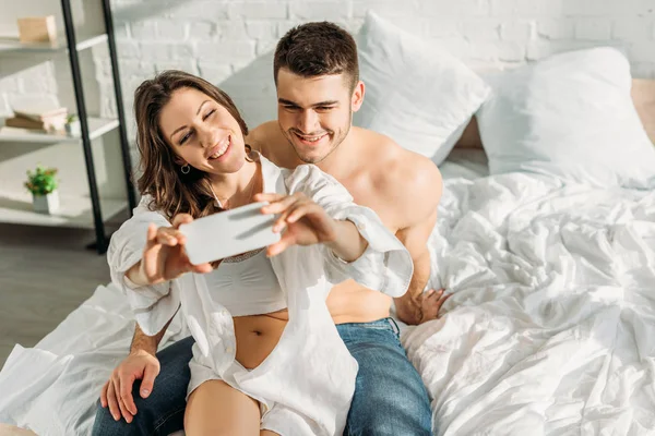 Chica feliz tomando selfie en el teléfono inteligente mientras está sentado en la cama con el novio sonriente - foto de stock