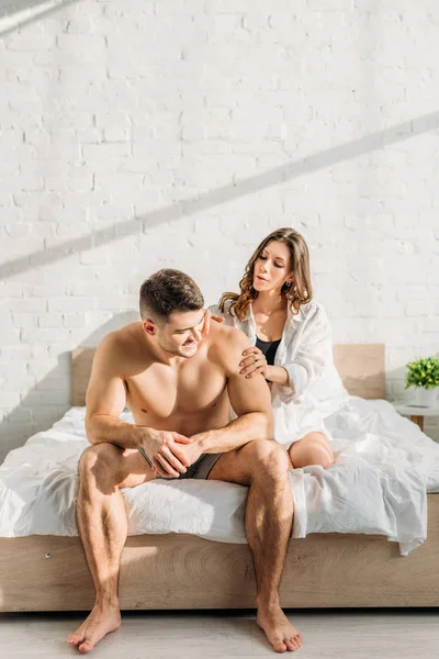 Приваблива дівчина в білій сорочці робить рукою еротичний масаж сексуальному хлопцеві, що сидить на ліжку — стокове фото