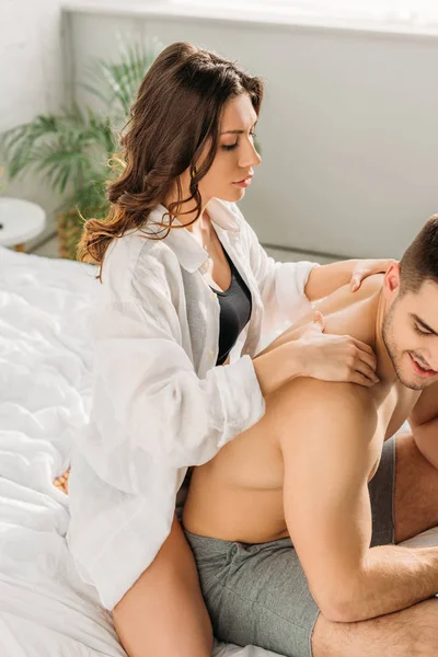 Sexy girl in white shirt making shoulders erotic massage to shirtless man sitting on bed — Stock Photo