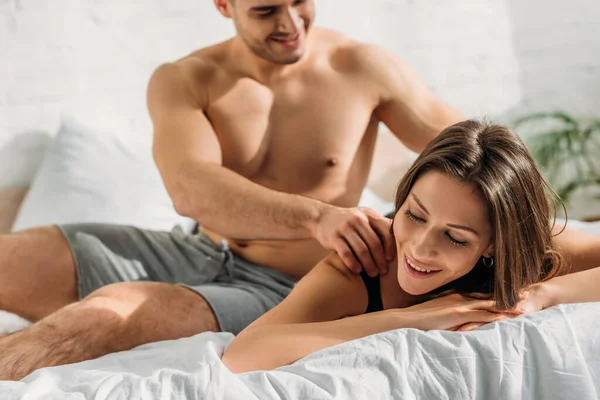 Selective focus of sexy shirtless man making shoulders erotic massage to pretty girlfriend lying and smiling with closed eyes — Stock Photo