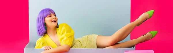 Tiro panorâmico de menina feliz bonita em peruca roxa como boneca deitada na caixa azul, isolado em rosa — Fotografia de Stock