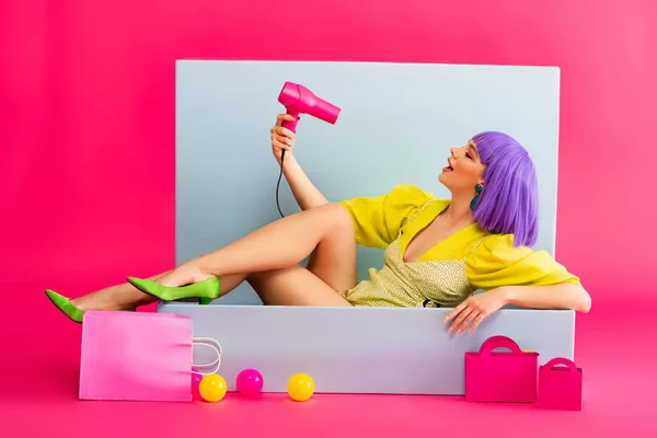 Chica feliz en peluca púrpura como muñeca usando secador de pelo mientras está sentado en la caja azul con pelotas y bolsas de compras, en rosa - foto de stock