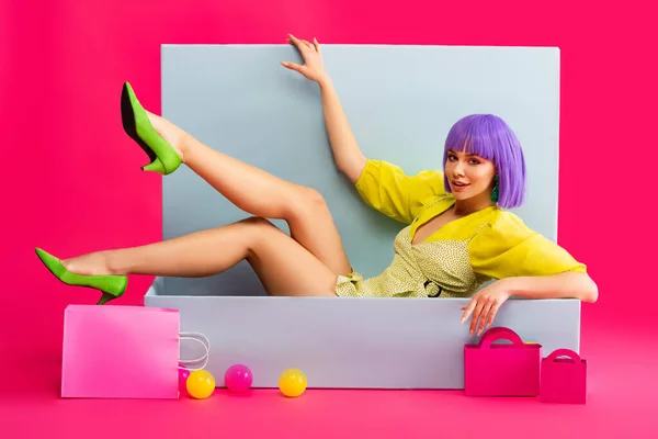 Bella ragazza emotiva in parrucca viola come bambola sdraiata in scatola blu con borse della spesa e palle, su rosa — Foto stock