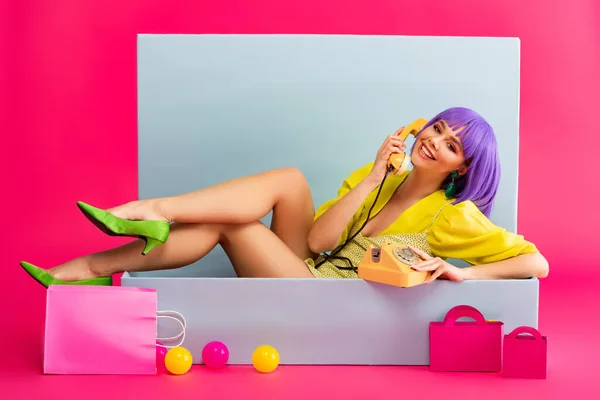 Smiling girl in purple wig as doll talking on vintage phone while sitting in blue box with balls and shopping bags, on pink — Stock Photo