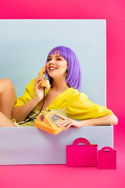 Bela menina sorridente em peruca roxa como boneca falando no telefone vintage enquanto sentado na caixa azul com sacos de compras, em rosa — Fotografia de Stock