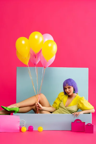Atractiva mujer en peluca púrpura como muñeca sosteniendo globos y sentado en caja azul con bolas y bolsas de compras, en rosa - foto de stock