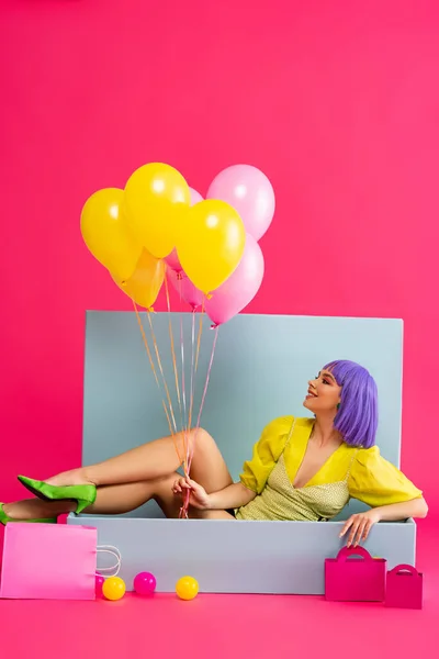 Happy girl in purple wig as doll holding balloons and sitting in blue box with balls and shopping bags, on pink — Stock Photo