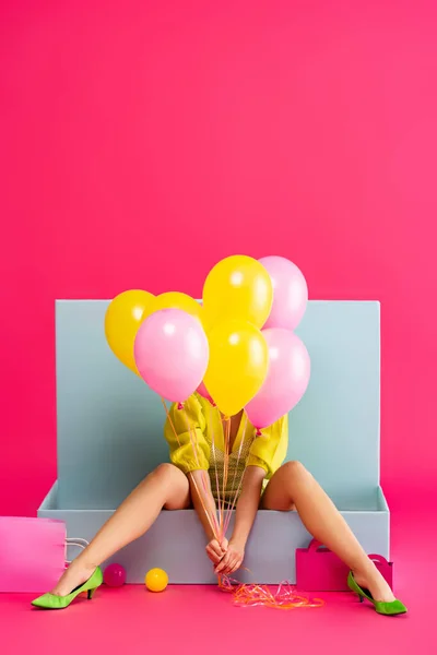 Jeune femme comme poupée tenant des ballons et assise dans une boîte bleue avec des boules et des sacs à provisions, sur rose — Photo de stock