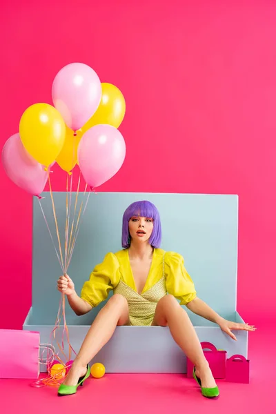 Menina emocional em peruca roxa como boneca segurando balões e sentado em caixa azul com bolas e sacos de compras, em rosa — Fotografia de Stock