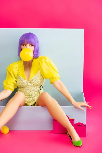 Pop art chica en peluca púrpura como muñeca que sopla goma de mascar mientras está sentado en la caja azul con bolsas de compras, en rosa - foto de stock