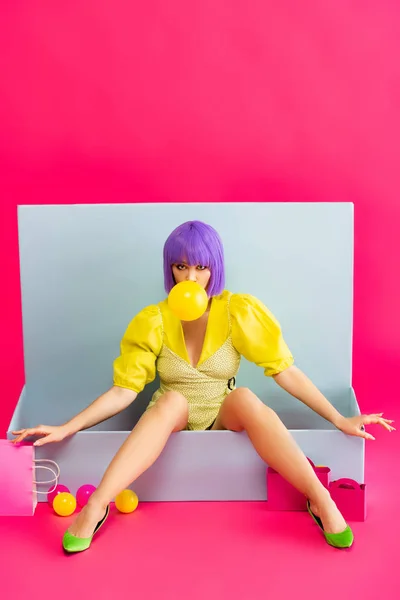Girl in purple wig as doll blowing bubble gum while sitting in blue box with balls and shopping bags, on pink — Stock Photo