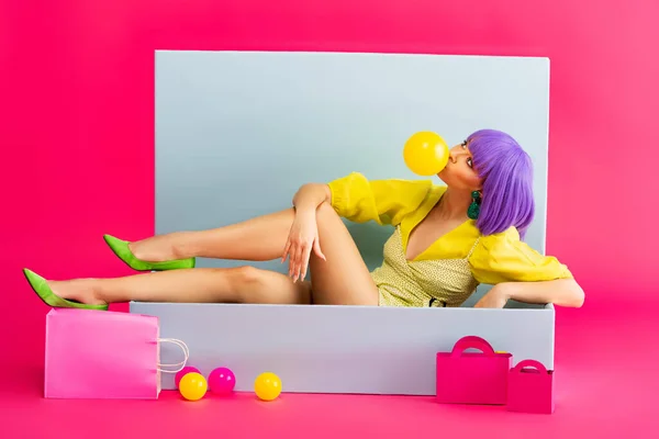 Pop art chica en peluca púrpura como muñeca que sopla goma de mascar mientras está sentado en la caja azul con bolas y bolsas de compras, en rosa - foto de stock